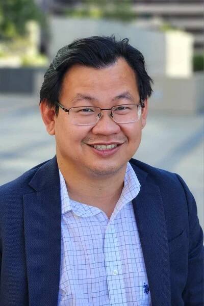 Professional portrait of an Asian professional in a navy suit and glasses smiling warmly against a blurred outdoor background.