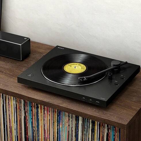Sony turntable playing a record, placed on a wooden console with a collection of vinyl records stored underneath, next to a black speaker