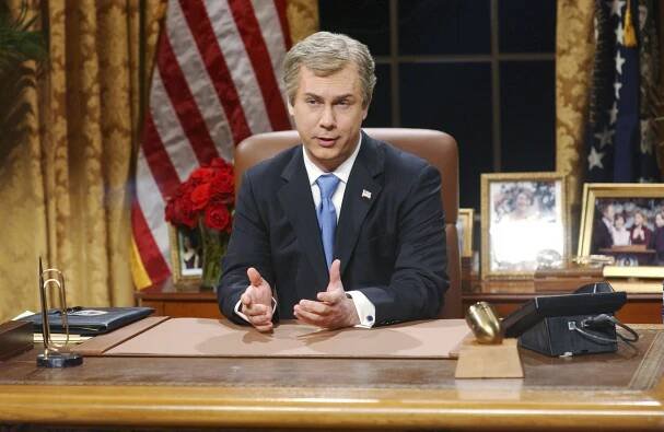 Chris Parnell as George W. Bush in a suit sits at a desk with an American flag and roses in the background