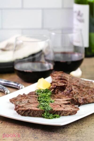 Steak gremolata on white platter with wine glasses in the background on a wood butcher block.