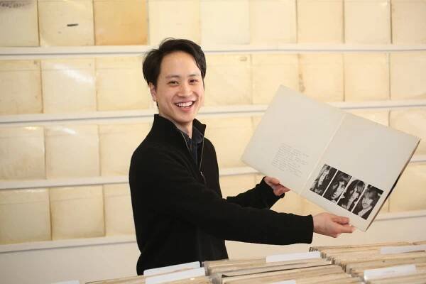 Rutherford Chang, a dark-haired young man wearing a black shirt, holds an open copy of the double LP “The White Album” in both hands. He stands in front of a wall displaying multiple copies of the album.