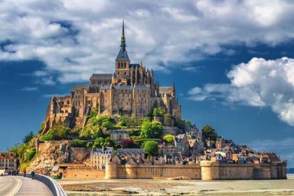 an abbey with a church spier on tp of a small hill with ccobalt blue sky and white clouds, Le Mont-Saint-Michel in France, Normandy
