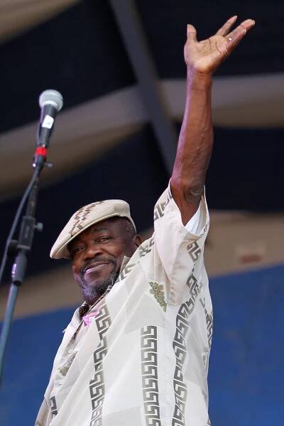 Clarence Henry, a bearded man wearing a patterned, white short-sleeved shirt and a cap, stands at a microphone on what looks like an outdoor stage and raises his left hand to wave to the crowd.