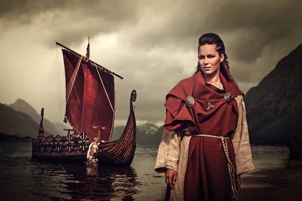 Vikingská žena s mečem a štítem, stojící poblíž Drakkar na břehu moře.