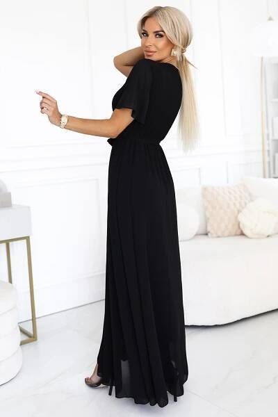 Modelka prezentuje elegancką czarną sukienkę maxi z szyfonu, z plisowanym dekoltem w kształcie litery V, krótkimi rękawkami i wiązaniem w talii; lekka i kobieca stylizacja marki Numoco Basic, idealna na wyjątkowe okazje.